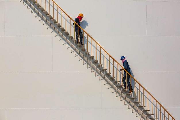 Работник нефтеперерабатывающего завода поднимается по металлической лестнице на промышленном резервуаре для хранения нефти