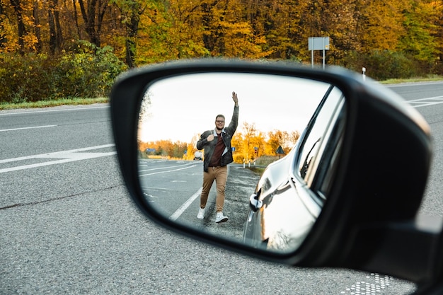 Reflected in the side mirror a man waves a hand at a car for the purpose of hitchhiking