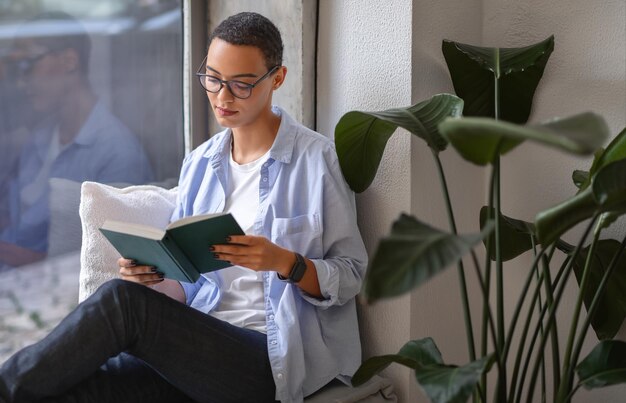 Reflecterende en ontspannen jonge vrouw die een boek leest bij het raam