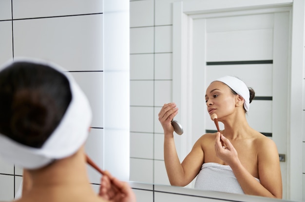 Reflectie in de spiegel van een jonge vrouw die een spiegel in de ene hand houdt en een markeerstift op haar gezicht aanbrengt