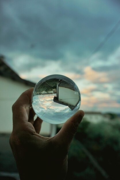 Foto reflectie van de hand van een persoon die een kristallen bol vasthoudt tegen de hemel