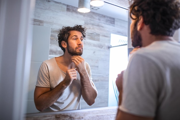 Photo reflection of handsome man with beard looking at mirror and touching face in bathroom grooming. man putting skincare facial treatment cream on face.