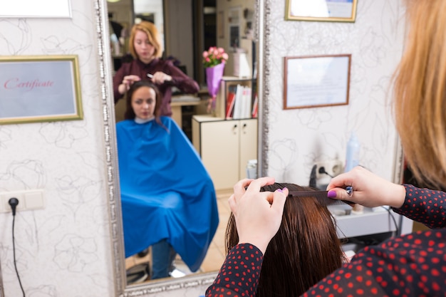 Reflection in Large Mirror of Young Blond Stylist Combing Hair of Brunette Client Wearing Blue Smock in Hair Salon