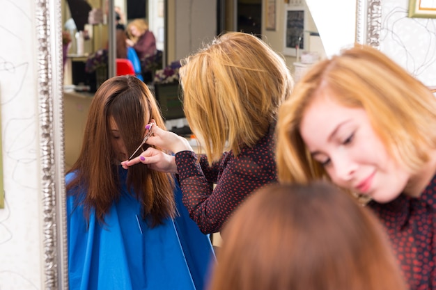 Riflessione nel grande specchio del salone di giovane stilista biondo che taglia i capelli di una cliente bruna - hair stylist che taglia la frangia per il cliente