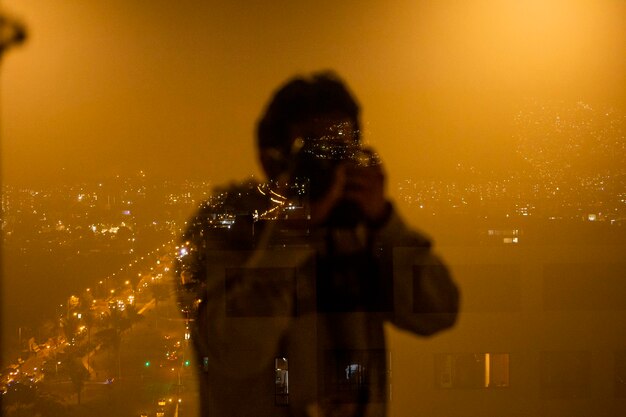 Reflection of man photographing on illuminated cityscape at night