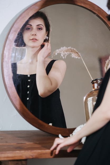 Reflection in a mirror of a cute sensual young woman in a black dress. Portrait of a beautiful brunette with short hair indoors.