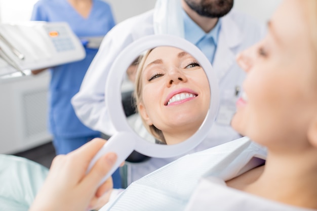 Reflection in mirror of healthy smile of pretty young smiling female patient of dental clinics after whitening teeth procedure by her dentist