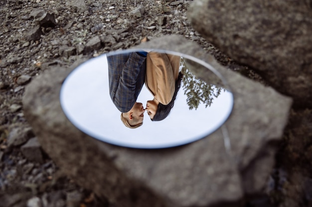 Reflection in the mirror of pair of woman and man who are closely tilted to each other The mirror lies on large stone.