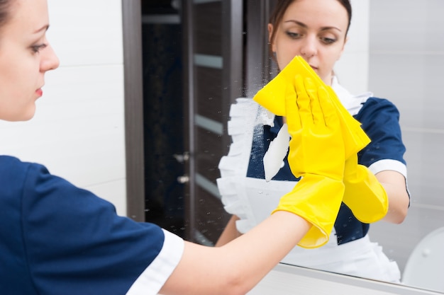 Отражение серьезной молодой взрослой горничной отеля, использующей желтую губку и резиновые перчатки, чтобы вытереть зеркало во время работы в ванной
