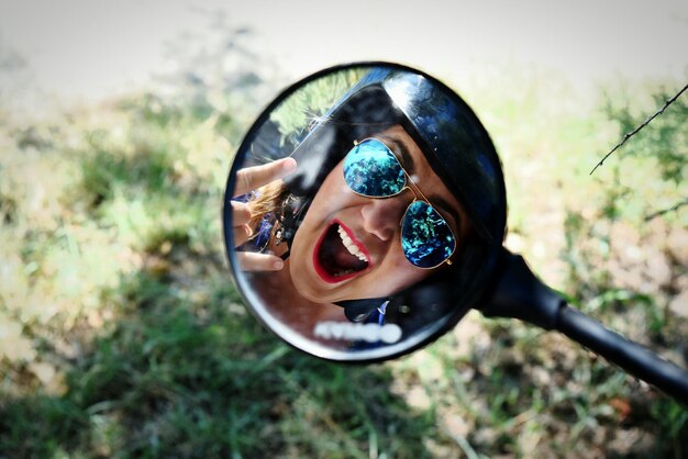 Photo reflection of woman wearing helmet and sunglasses gesturing horn sign in side-view mirror of motorcycle