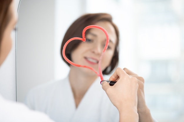 Reflection of young brunette female in white bathrobe drawing heart with crimson lipstick on mirror