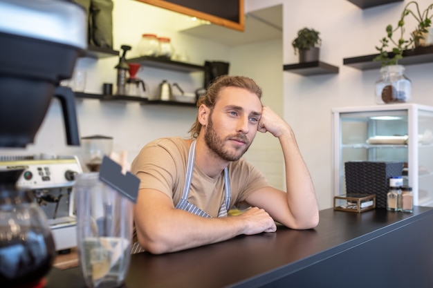 Reflections. Pensive calm young man in apron standing behind bar with his head on hand looking into distance