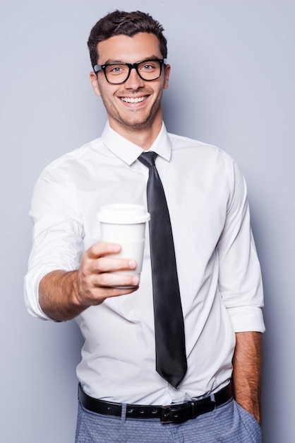 心をリフレッシュしてください！シャツとネクタイで自信を持って若い男がコーヒーカップを伸ばして、灰色の背景に立って笑っている
