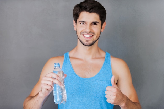 Refreshing after training. Cheerful young muscular man holding a bottle with water and thumb up while standing against grey background