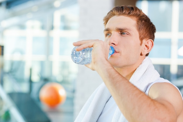 운동 후 상쾌함. 체육관에 서 있는 동안 어깨에 수건을 들고 물을 마시는 피곤한 청년