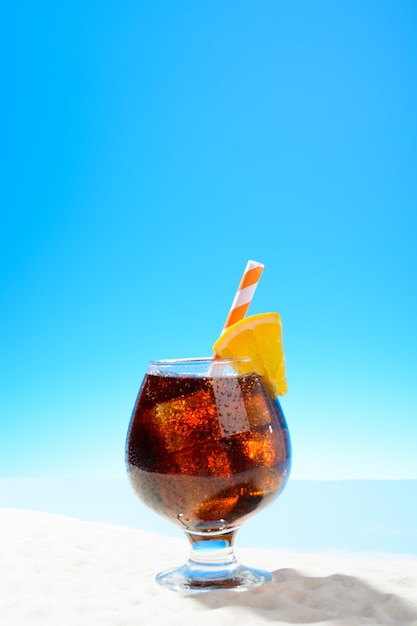 Refreshing Cola with ice and a slice of orange in a glass on the sandy coast