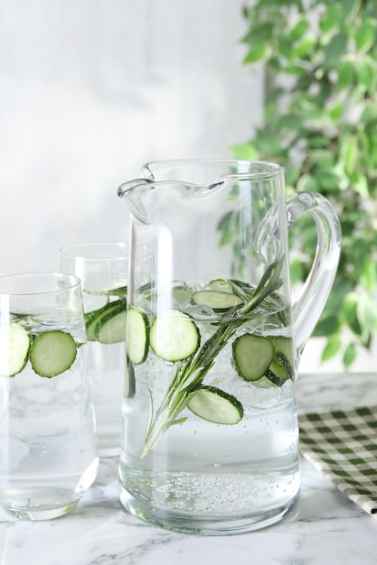 Refreshing cucumber water with rosemary in jug and glasses on white marble table