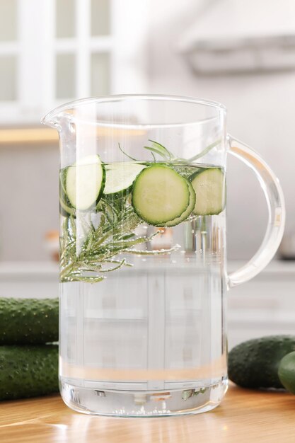 Refreshing cucumber water with rosemary in jug and vegetable on wooden table closeup