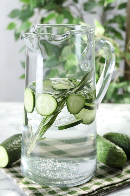 Refreshing cucumber water with rosemary in jug and vegetables on table closeup