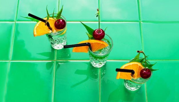 Refreshing drinks in crystal glasses leaves cherry orange slice green tile background shadow Sweet cold natural cocktail