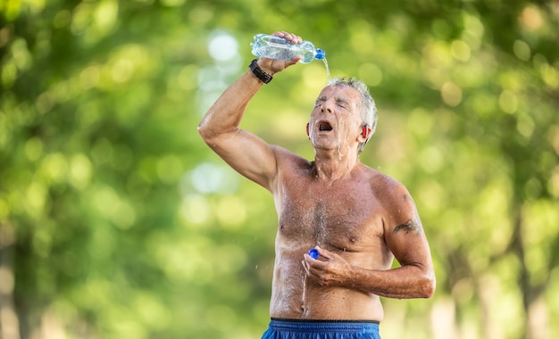 Refreshing elderly man pours water over him in the green forest to cool down on a hot sunny day.