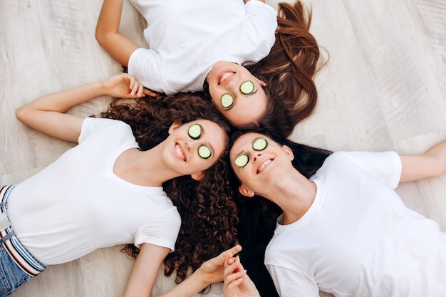 Refreshing eye mask. Beautiful smiling girls and their mother lie on the floor, make a face mask of cucumbers