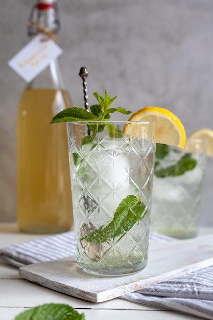 Освежающий лимонад с ледяным лимоном и мятой с бутылкой сиропа на светлой поверхности фона