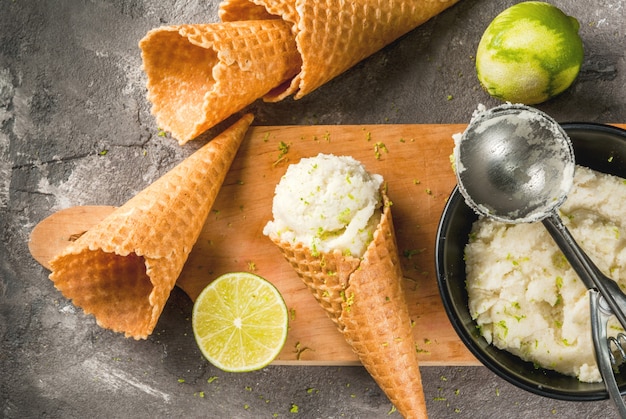 Refreshing lime sorbet in a bowl with a spoon for ice cream. With horns for ice cream (one full of ice cream), limes, grater for peel. On an old gray concrete table. Top view copyspace