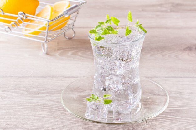 Refreshing mineral water with ice cubes and mint leaves in a transparent glass and lemon in a basket on a wooden table