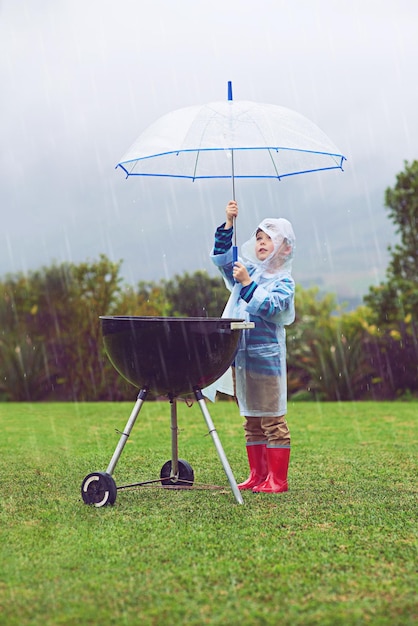 Regenparaplu en een jongen die een barbecue buiten op een veld bedekt terwijl hij alleen eten kookt in de winter Regenjaskinderen en verzekeringen met een jong mannelijk kind dat een bbq probeert te beschermen tijdens stormachtig weer