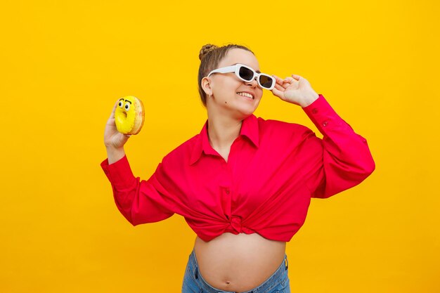 Regnant woman eats sweet donuts on a yellow background a pregnant woman in a pink shirt and glasses eats a sweet cake harmful food during pregnancy