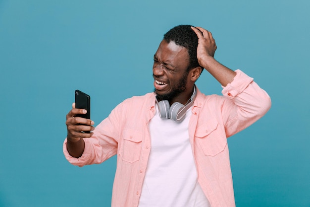 Regret grabbed head holding phone young africanamerican guy wearing headphones on neck isolated on blue background