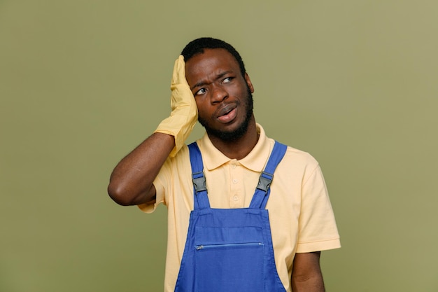 Regret grabbed head young africanamerican cleaner male in uniform with gloves isolated on green background