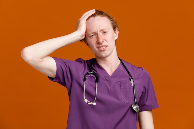 Regret grabbed head young male doctor wearing uniform with stethoscope isolated on orange background