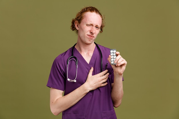 Regret putting hand on heart young male doctor wearing uniform with stethoscope holding pills isolated on green background