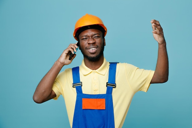 Regret young african american builder in uniform speaks on the phone isolated on blue background