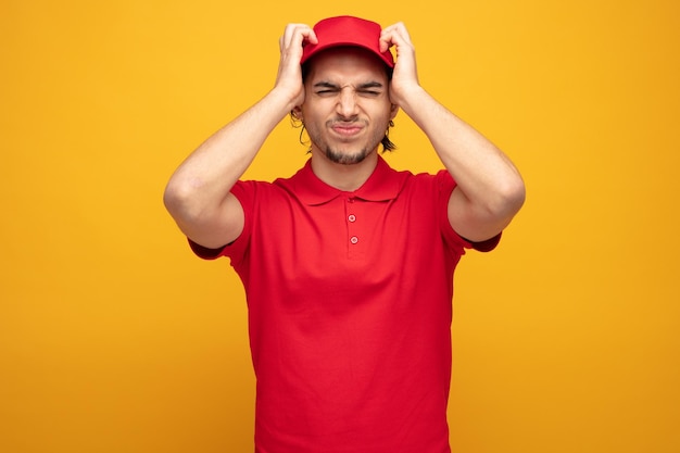 Giovane uomo di consegna dispiaciuto che indossa l'uniforme e il cappuccio che tiene le mani sulla testa con gli occhi chiusi isolati su sfondo giallo