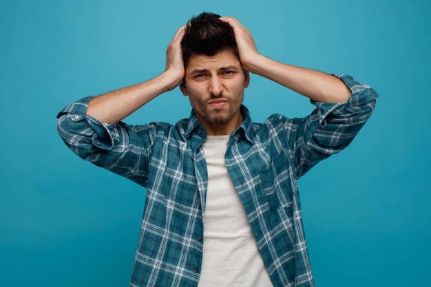 Regretful young man keeping hands on head looking at camera isolated on blue background