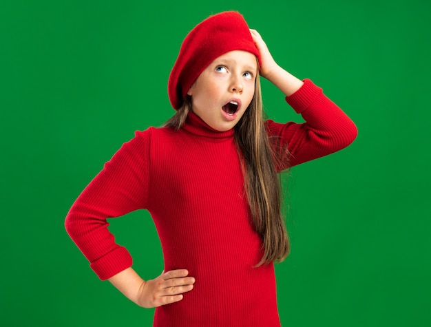 Regretting little blonde girl wearing red beret looking up keeping hand on head and on waist with open mouth isolated on green wall