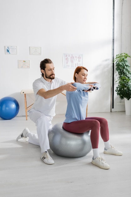 rehabilitation specialist assisting woman sitting on fitness ball and exercising with dumbbells