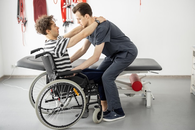 Rehabilitation specialist helps man to get up from a wheelchair at clinic