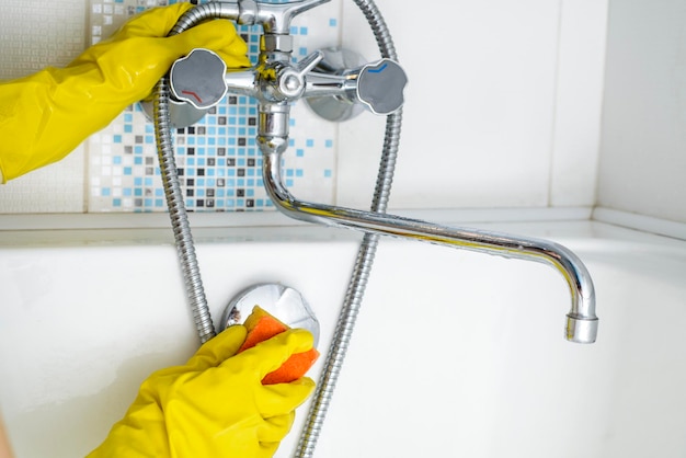 Reiniging van de gootsteen en kraan in de badkamer met wasmiddel in gele rubberen handschoenen met oranje spon