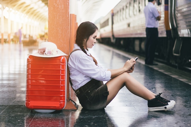 Reis Aziatische zwangere de vrouw bekijkt Smartphone met een rode koffer bij station.