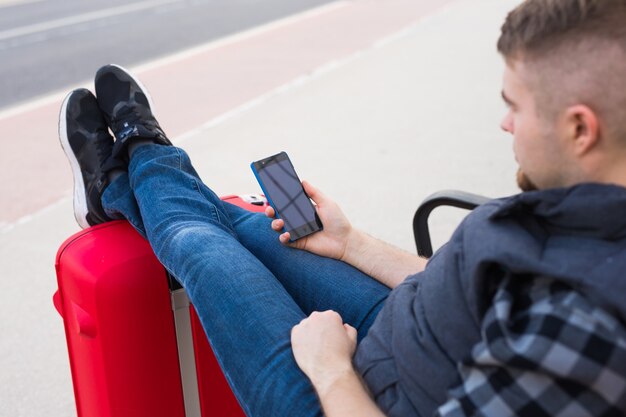 Reis-, toerisme- en mensenconcept - knappe man zit en zette zijn voeten op koffer, kijkend naar iets in zijn mobiel.