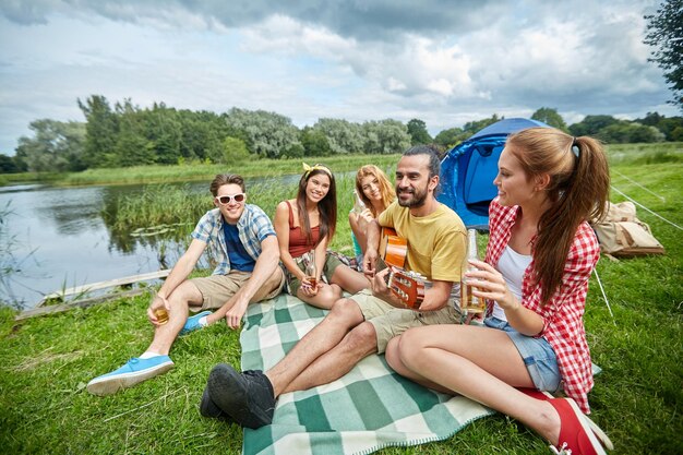 Reis, toerisme, stijging, picknick en mensenconcept - groep gelukkige vrienden met tent en dranken die gitaar spelen bij het kamperen