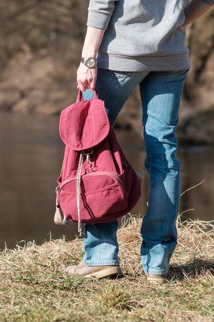 Reizen concept. vrouw aan de oever van de rivier gekleed in spijkerbroek en grijze hoody, rode rugzak houden en ver weg kijken. lente seizoen.