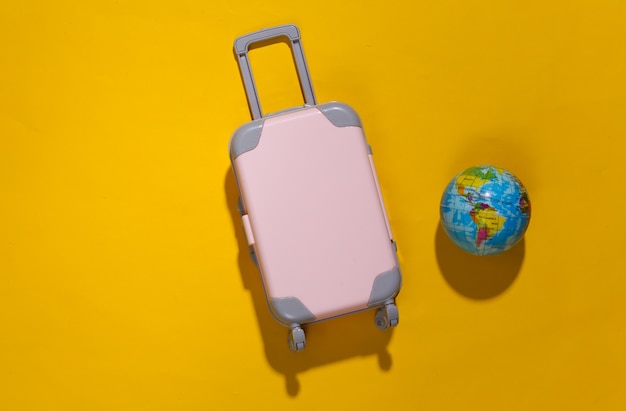 Reizen minimalisme. Mini plastic reiskoffer en globe op gele achtergrond met diepe schaduw. Minimale stijl. Bovenaanzicht, plat gelegd