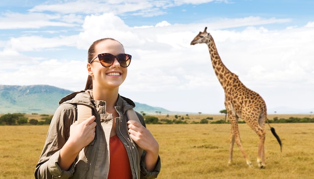 reizen, toerisme en mensenconcept - gelukkige jonge vrouw in zonnebril met rugzak over afrikaanse savanne en girafachtergrond