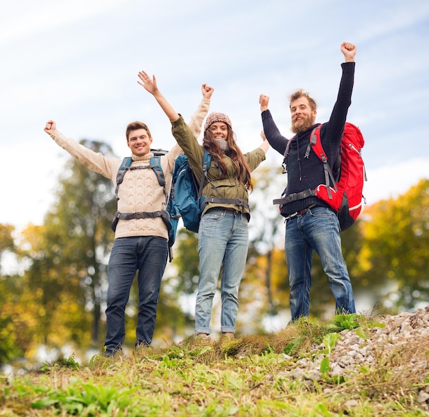 reizen, toerisme, wandeling, gebaar en mensen concept - groep lachende vrienden met rugzakken die handen opsteken over natuurlijke achtergrond