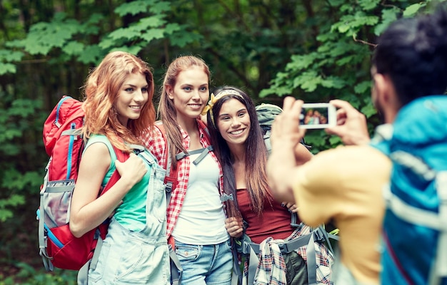 reizen, toerisme, wandeling, technologie en mensenconcept - groep lachende vrienden die met rugzakken lopen en foto's maken met een smartphone in het bos